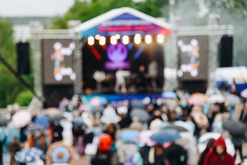 music festival in city park, crowd of people backview, rock group performing on stage, green trees and summer day, blurred background