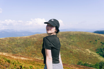 A woman enjoying a hike on a mountain trail smiles while surrounded by lush hills and vibrant landscapes under a clear blue sky during a sunny day.