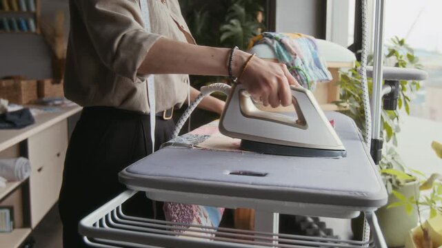 Medium shot of hands and midsection of anonymous seamstress ironing out creases on sewn patchwork piece, while creating new quilt design in artisan workshop