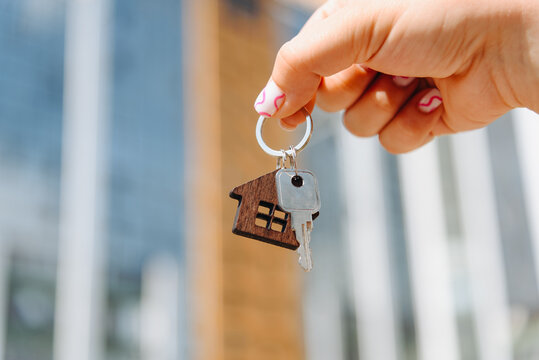 woman hand with metal key, keychain in shape of private house, modern brown brick multi-storey apartment building in background, purchase of real estate, real estate services