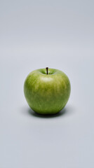 A vibrant green apple sits alone in the center of a light gray background, highlighting its fresh texture and natural gloss
