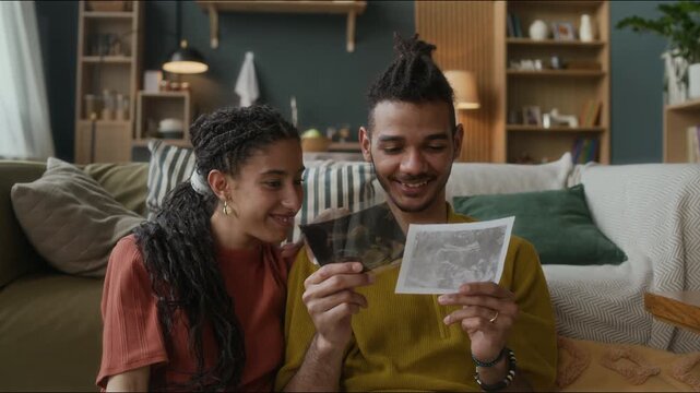 Medium shot of happy young biracial parents to be looking at baby ultrasound pictures sitting in cozy modern living room, chatting and feeling excited to meet their little one - Powered by Adobe