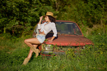 Beautiful woman with a straw hat poses next to an old abandoned car surrounded by vegetation.