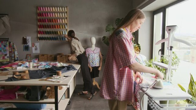 Medium full shot of busy Caucasian seamstress pressing unfinished patchwork fabric piece using professional steam iron system, while female colleague is preparing sewing tools on cutting table