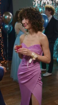 Vertical shot of young girl in pink fancy evening dress holding glass of punch while talking to unrecognizble classmate at high schol prom party