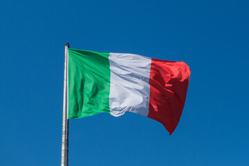 The Italian tricolor flag, with its green, white, and red stripes, waves gracefully in the wind against a clear and bright blue sky.