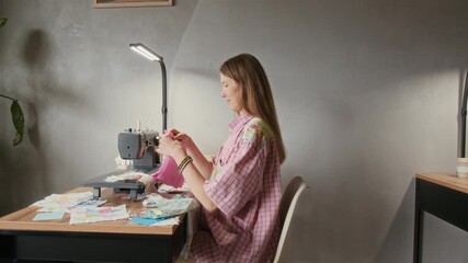 Medium tracking shot of smling junior seamstress pasing over piece of textile arment to senior female coleague, who is moving overto sewing machine and prepring to stitch unfinished arts