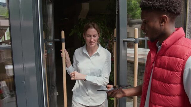 Young African American courier in red sleeveless vest knocking on door and giving bouquet of daises to woman