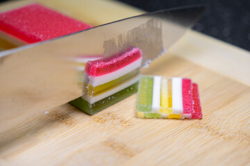 A sharp kitchen knife precisely slicing vibrant multi-layered jelly candy block on a bamboo cutting...