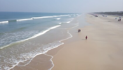 A wide sandy beach with gentle waves rolling in, and a few people walking along the shore on a sunny day.