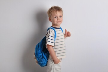 Young boy with a cheerful expression showing a thumbs up while wearing a striped shirt and a blue backpack on white background.