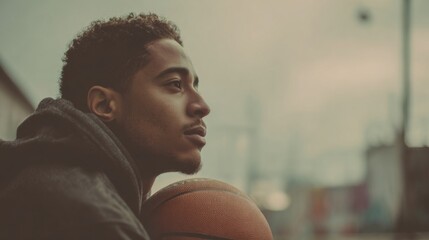 Young African American man sitting on indoor basketball court holding basketball and looking thoughtful.