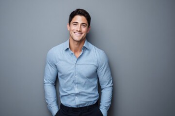 Smiling young man in light blue shirt standing against plain gray background.