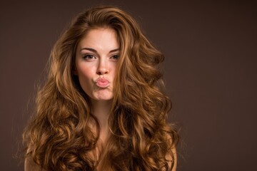 Young woman with long curly red hair making kissing face against plain brown background.