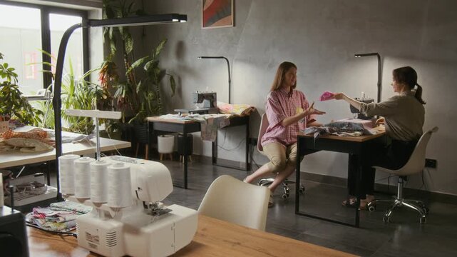 Wide shot of artisan sewin workshop, with professionl serger machine on workto in foreground, and young aucasian seamstress passin over piece of fabric to clleague for sewing