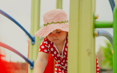 A young child wearing a red polka dot dress and a hat is climbing on colorful playground equipment...