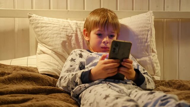 Cute kid boy in pajamas lying in bed watching smartphone, smiling. Children's screen time. Child plays games using phone at the evening or night. Social media addiction. Childhood, modern technology
