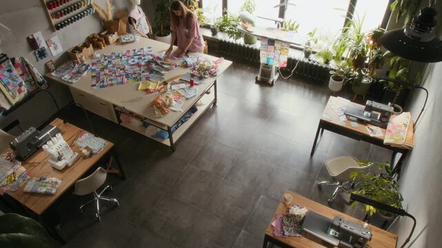 Full high surveillance time lapse shot of two young female seamstresses working together in sewing workshop, discussing patchwork designs by cutting table, operating professional machines