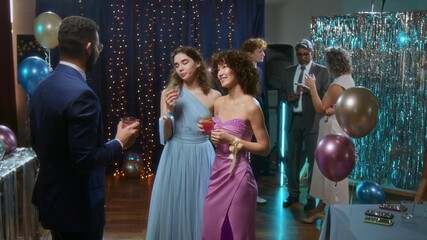 Young man in navy blue suit talking to two girls in elegant evening dresses and drinking punch at high school prom party