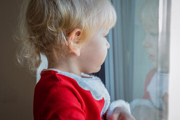 Blond little boy in Santa outfit looking through window with reflection during Christmas