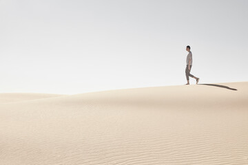 Man walking barefoot on sandy dunes under bright sky.