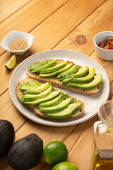 Avocado slices laying on toasted bread on a white dish. It sits on a wooden table with ingredients like sesame seeds, avocadoes, lime, oil and dried tomatoes.