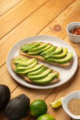 Avocado slices laying on toasted bread on a white dish. It sits on a wooden table with ingredients like sesame seeds, avocadoes, lime, oil and dried tomatoes.