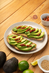 Avocado slices  laying on toasted bread with dried tomato on a white dish. It sits on a wooden table with ingredients like sesame seeds, avocadoes, lime, oil and dried tomatoes.
