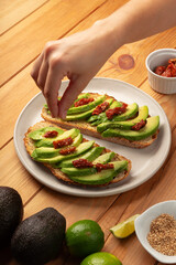 Hand serving sesame seeds on toasted bread with avocado slices and dried tomato. They lay on a white dish on a wooden table with bowls with ingredients.