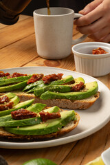 Avocado and dried tomato toasts on a white dish sitting on a wooden table. A hand is serving coffe...