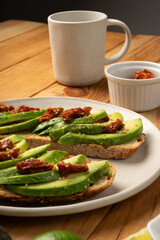 Avocado and dried tomato toasts on a white dish sitting on a wooden table with a mug of coffe in...