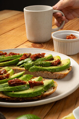 Avocado and dried tomato toasts on a white dish sitting on a wooden table. A hand is grabing a...