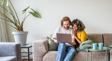 Fototapeta na wymiar Multiracial young couple watching computer laptop sitting on the sofa at home - Happy diverse husband and wife using pc online services - Technology life style concept