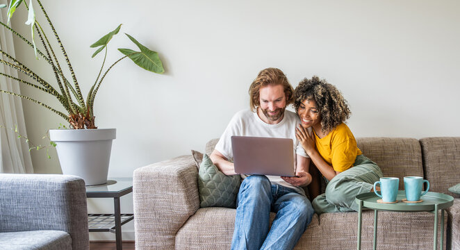 Multiracial young couple watching computer laptop sitting on the sofa at home - Happy diverse husband and wife using pc online services - Technology life style concept
