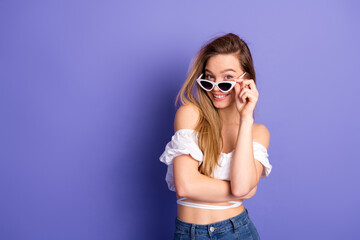 Charming young woman in white top and sunglass posing against a violet background with a cheerful...