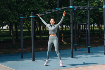young slim caucasian woman doing jumping jack exercises outdoors in summer, street sports ground, grey sportswear, green trees in background, healthy lifestyle concept
