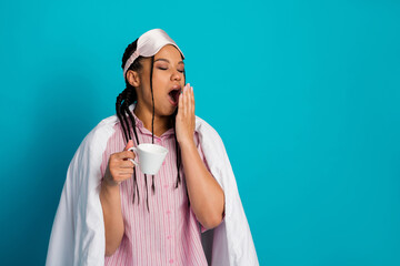 Tired young woman in pink pajamas yawning while holding a white cup against a bright blue background