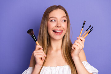 Smiling young woman holding makeup brushes in a joyful pose against a purple background