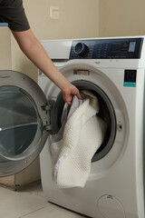 A person's arm reaches into a front-loading washing machine to put in a light-colored, quilted...