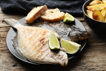 Tasty roasted flounder fish served with potatoes, bread and lime slices on wooden table, closeup. Homemade seafood dish