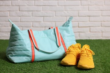Stylish sports bag and sneakers on green grass against white brick wall, closeup
