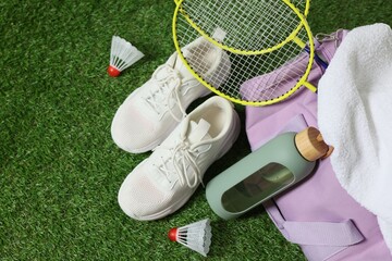 Stylish sports bag with badminton rackets, shuttlecocks, bottle and sneakers on green grass, flat...