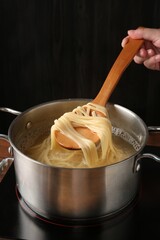 Woman taking boiled tagliatelle pasta with spoon from pot on electric stove, closeup