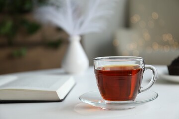 Cup of tea and book on white table against blurred lights, closeup