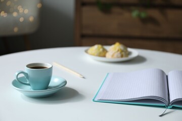 Notebook, cup of coffee and cookies on white table against blurred lights