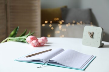 Notebook with pencil, alarm clock and tulips on white table against blurred lights