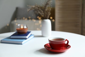 Cup of tea, books and burning candle on white table against blurred lights