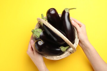 Woman with wicker basket of fresh eggplants on yellow background, top view