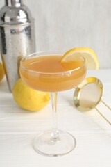 Martini cocktail with lemon slice in glass, fruit and shaker on white wooden table, closeup
