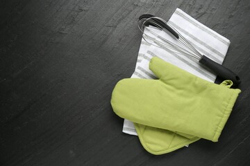 Green oven glove, towel, potholder and whisk on dark textured table, flat lay. Space for text